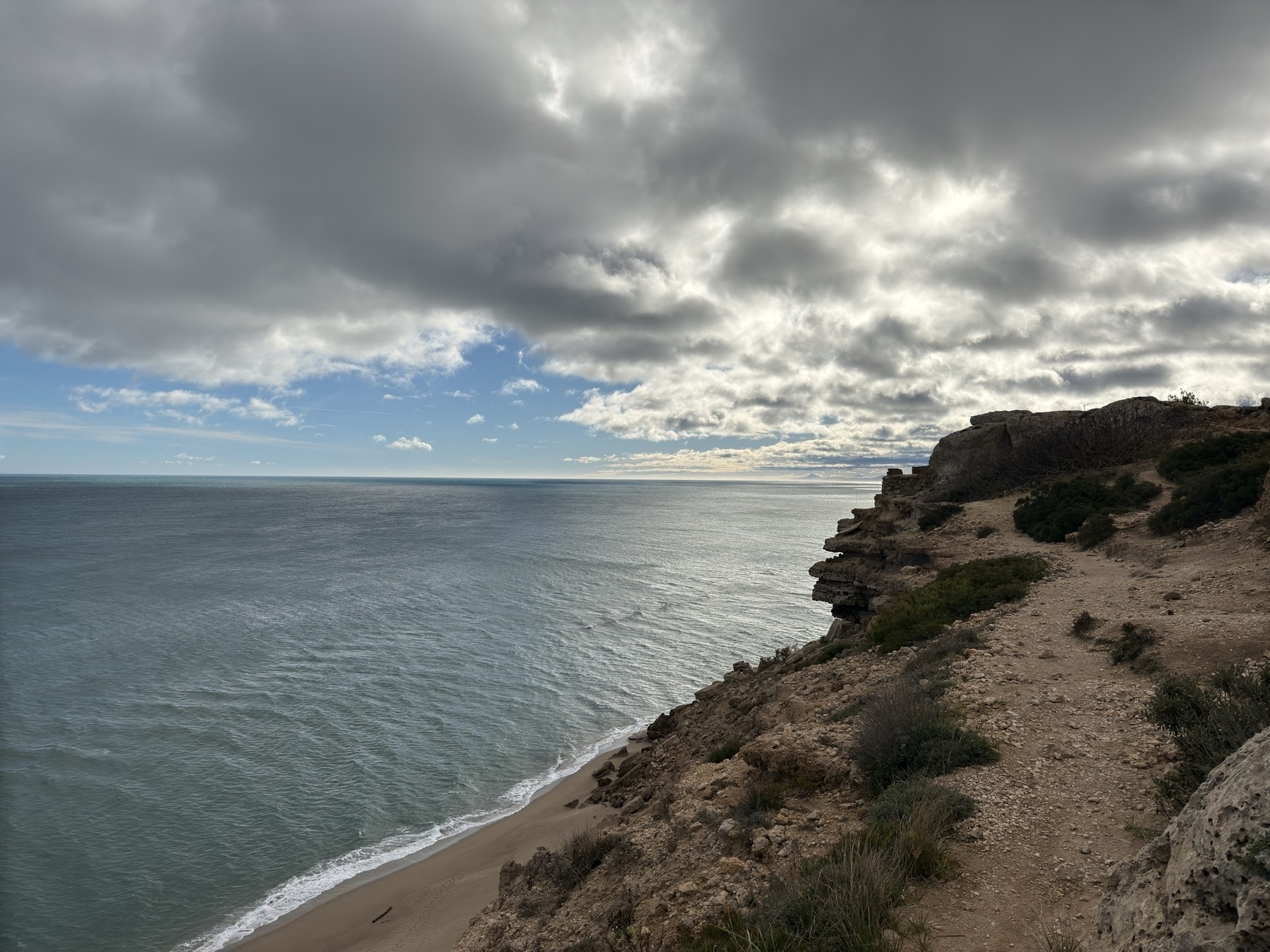 Photo of a shoreline. In the foreground is the sandy cliff the photographer is standing on, and below it the sandy beach. At the shoreline there is white foam leading into a dark blue sea (the Mediterranean). A sliver of blue sky on the horizon is crowded out by steel-grey clouds, backlit by the sun.
