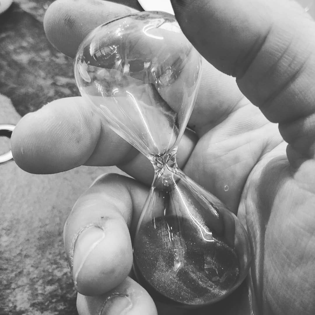 Auto-generated description: A close-up black and white photo of a hand holding a small, clear hourglass with sand inside.