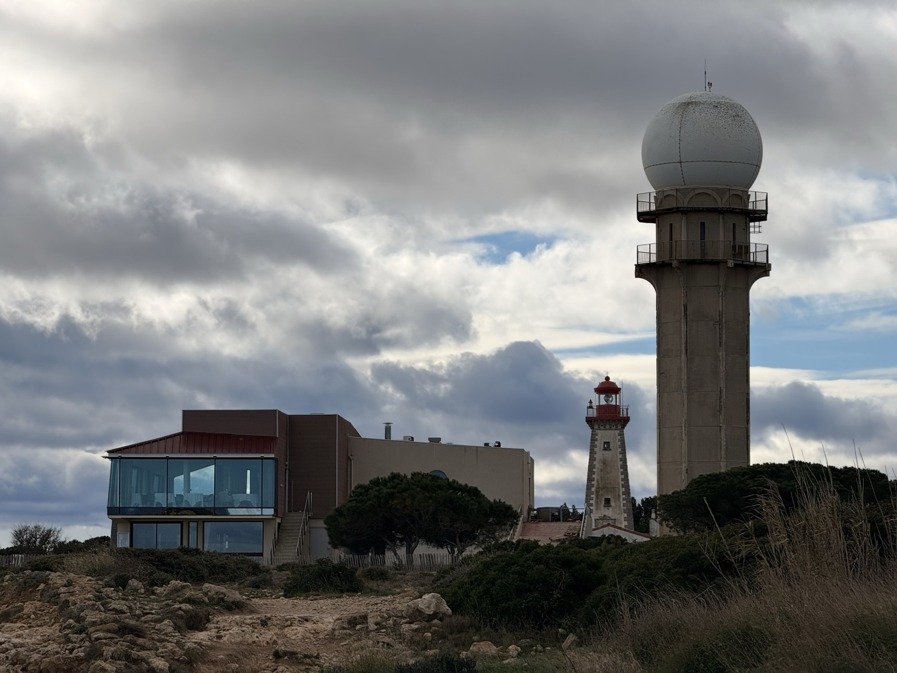A modern radar tower stands beside a traditional lighthouse, set against a cloudy sky. Next to both of them is a building containing a restaurant, with a huge panoramic glass window reflecting the blue sky.