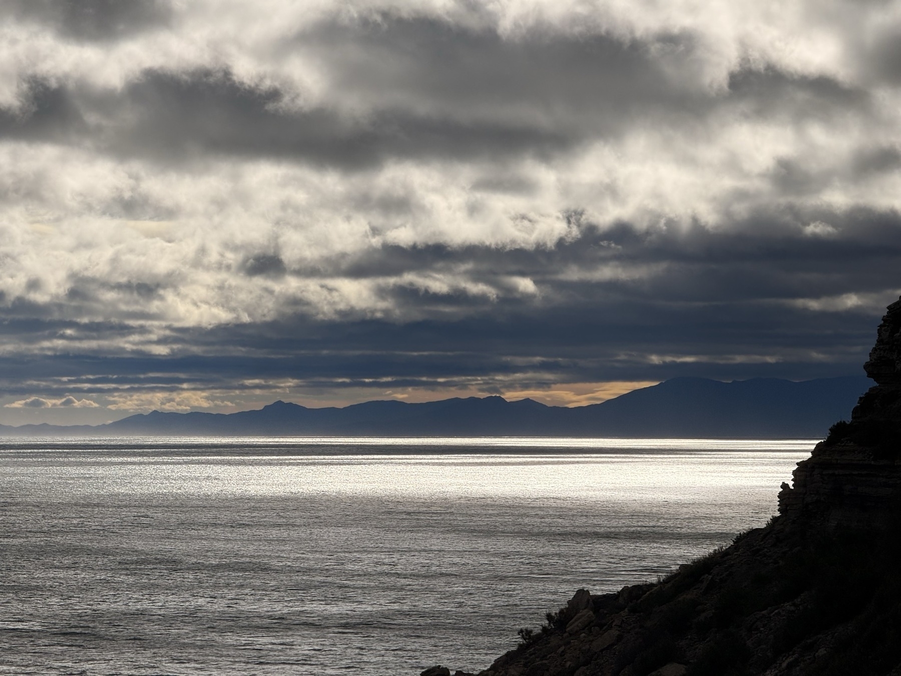 Dark clouds hover over a tranquil sea with distant mountains silhouetted on the horizon.