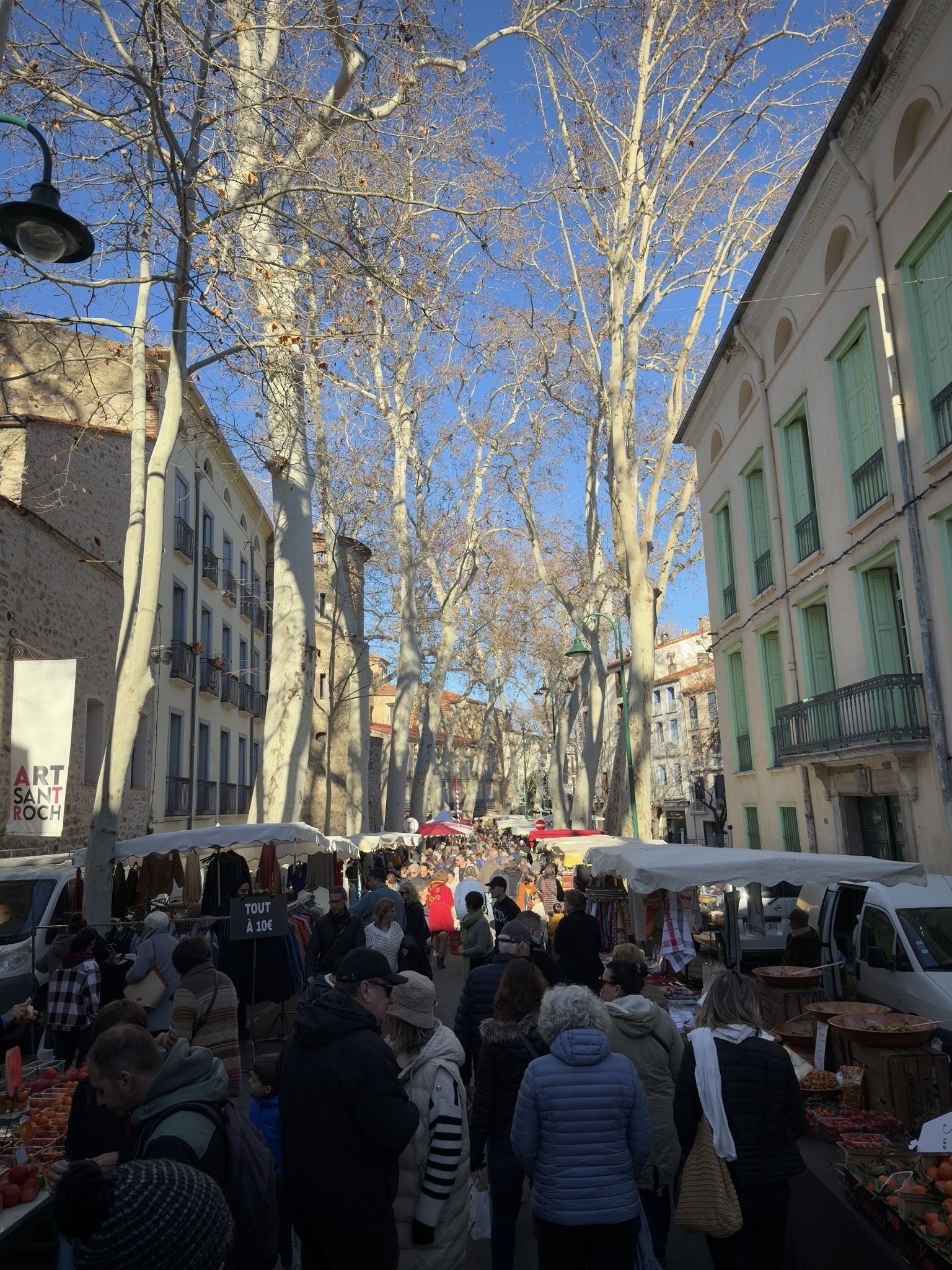 Photograph of a street lined with sun kissed trees. Many people are walking between the market stalls that line the street.