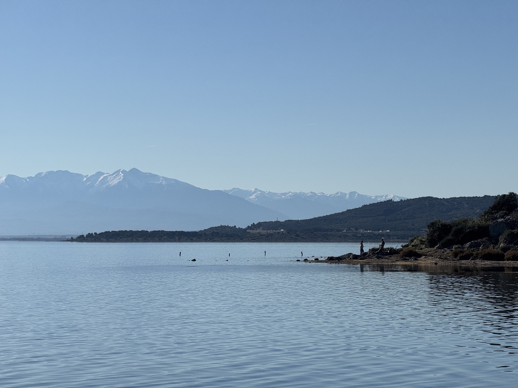 A serene landscape features calm water in the foreground, a distant mountain range, and a clear blue sky.
