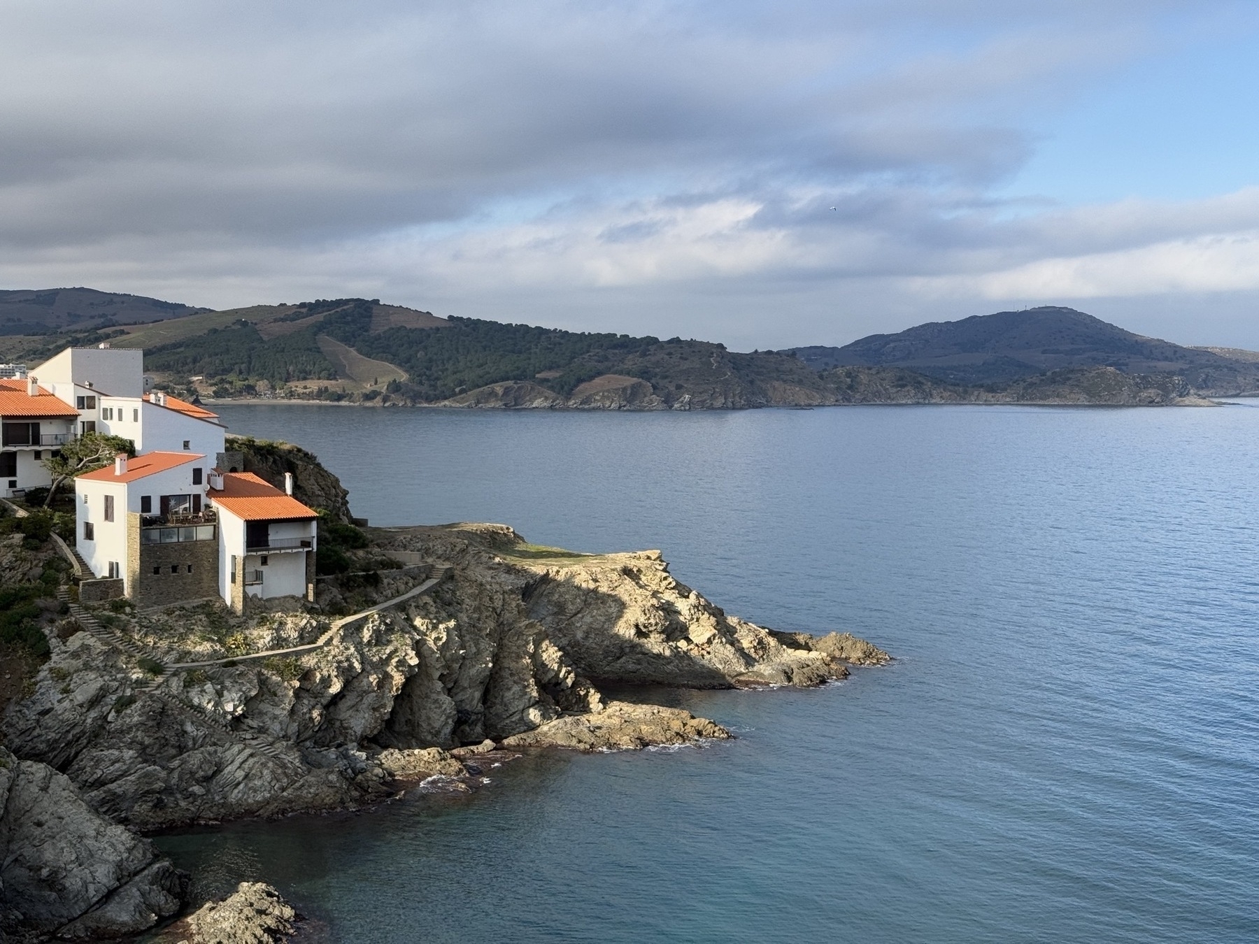 Coastal houses with red roofs are perched on rocky cliffs overlooking a calm sea, with hills in the background under a partly cloudy sky.