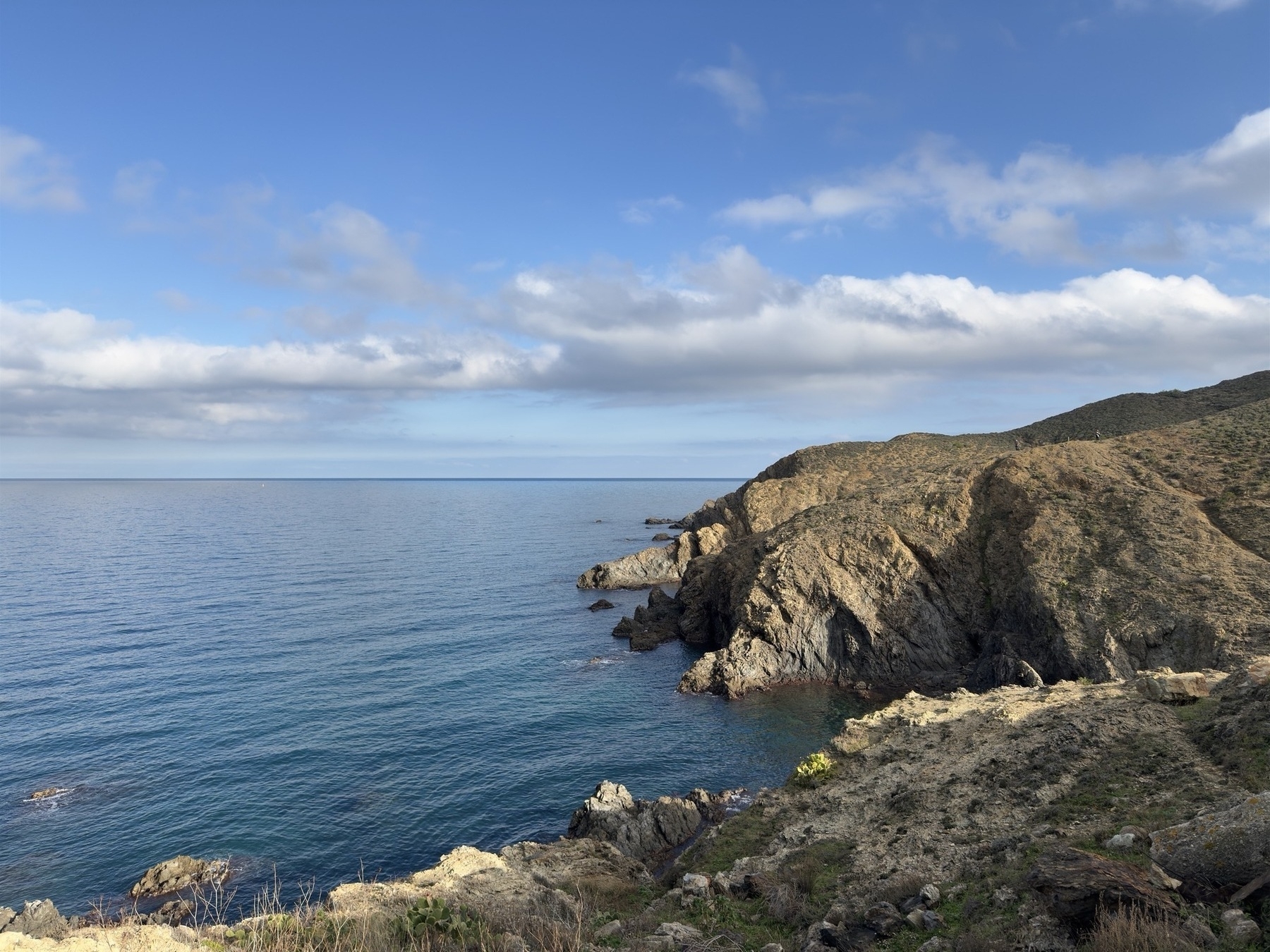 A rocky coastline meets a calm, expansive sea under a partly cloudy blue sky.