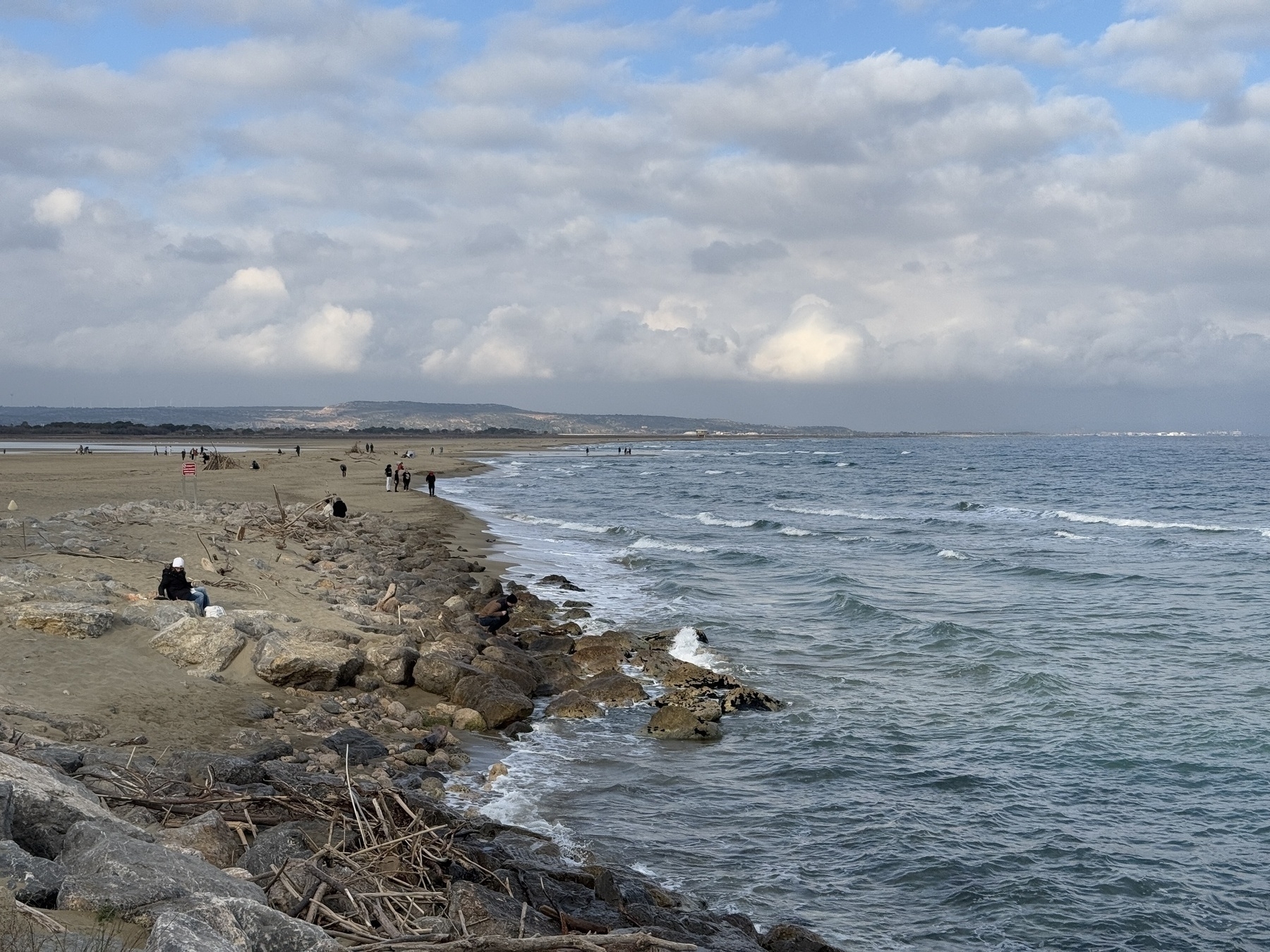 Photo: A rocky shoreline with waves gently crashing under a partly cloudy sky, with a few people walking along the beach.